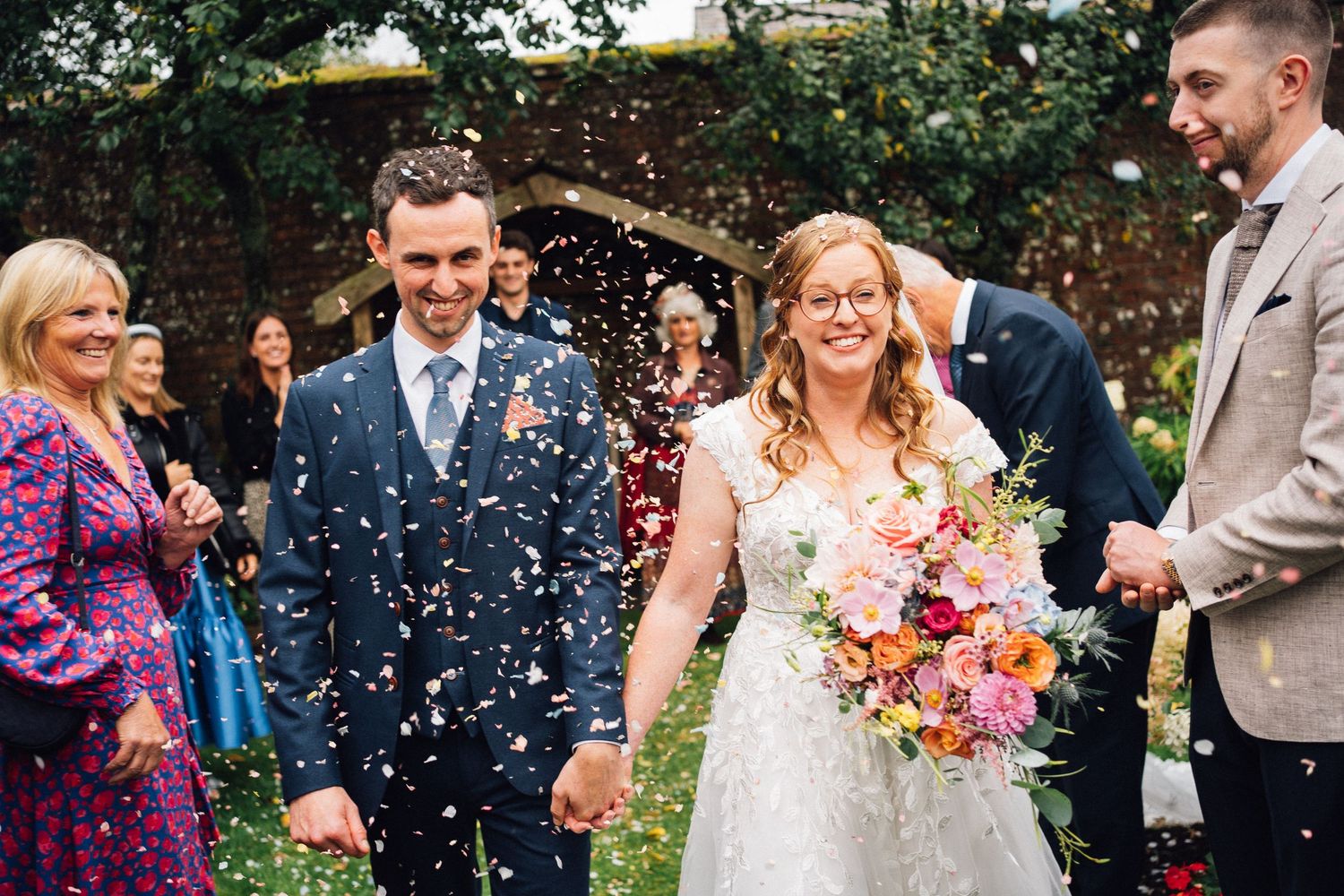 Wedding guests celebrate by throwing colorful confetti in an outdoor garden setting during a joyful celebration.