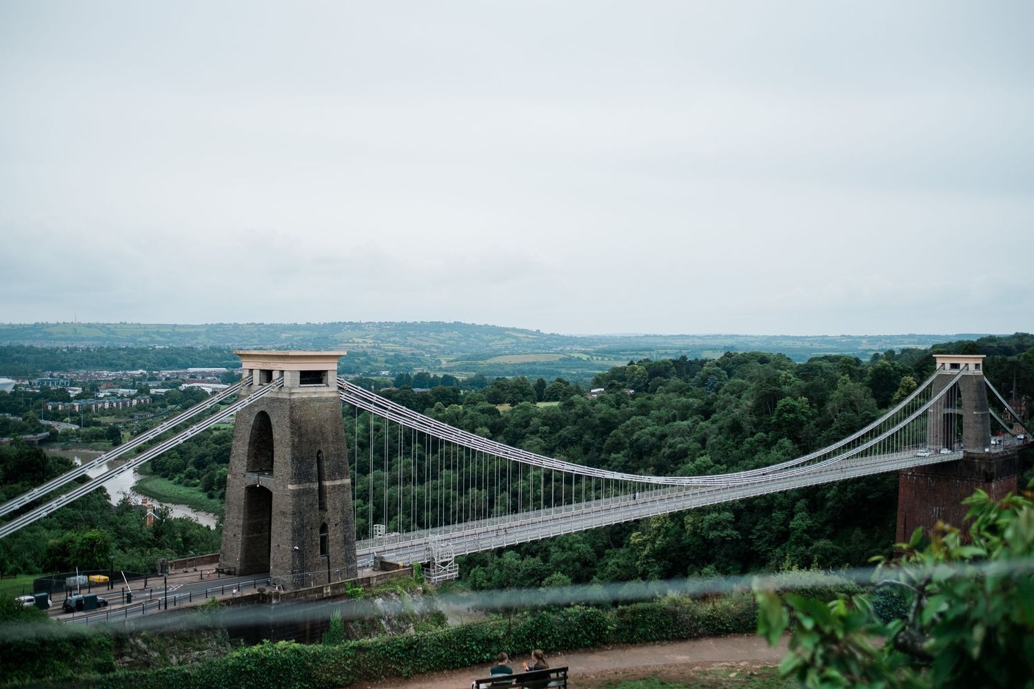 Clifton Suspension Bridge spanning a lush green gorge in Bristol, England, under a cloudy sky with rolling hills beyond.