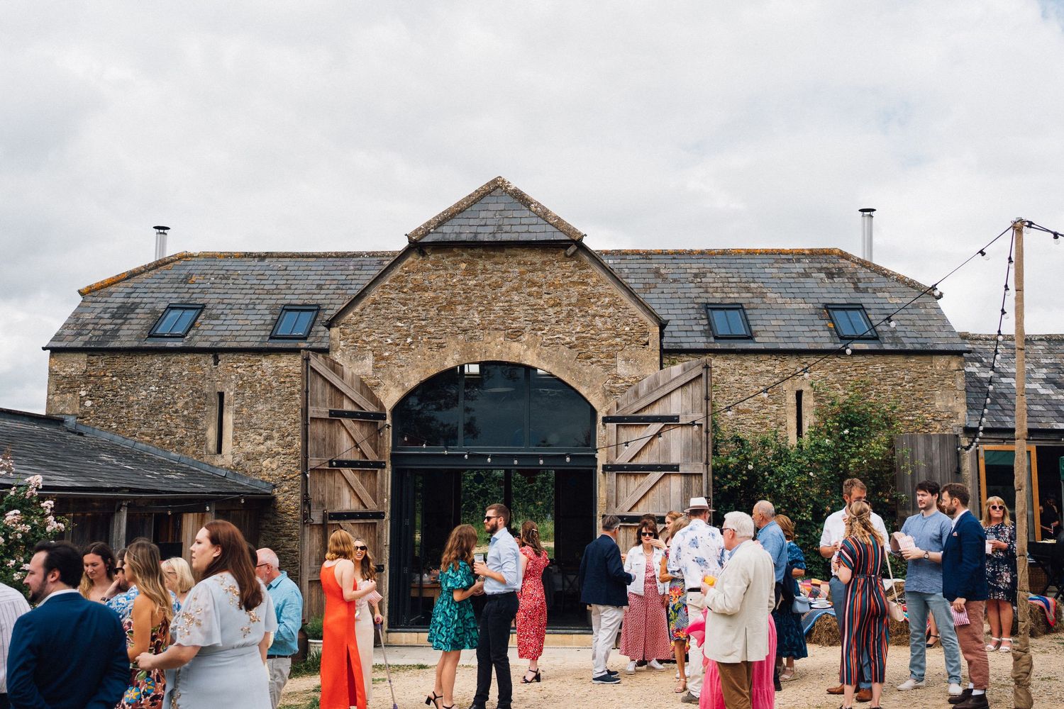 Guests mingling outside a rustic stone barn wedding venue under cloudy skies with string lights and wooden doors.