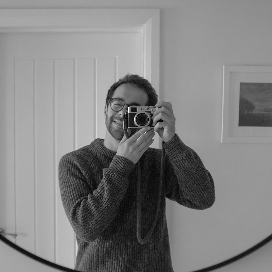 Black and white mirror selfie of a person holding a vintage film camera, wearing a knit sweater, indoors.