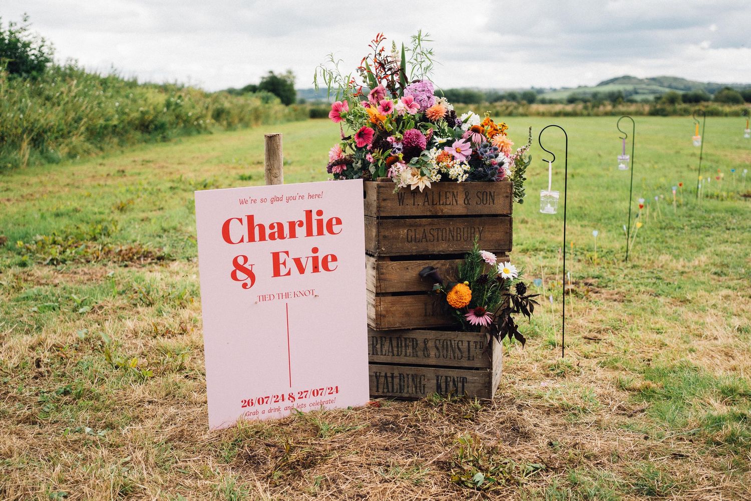 Rustic wooden crates and pink signage decorate a grassy outdoor wedding venue with rolling hills in the background.