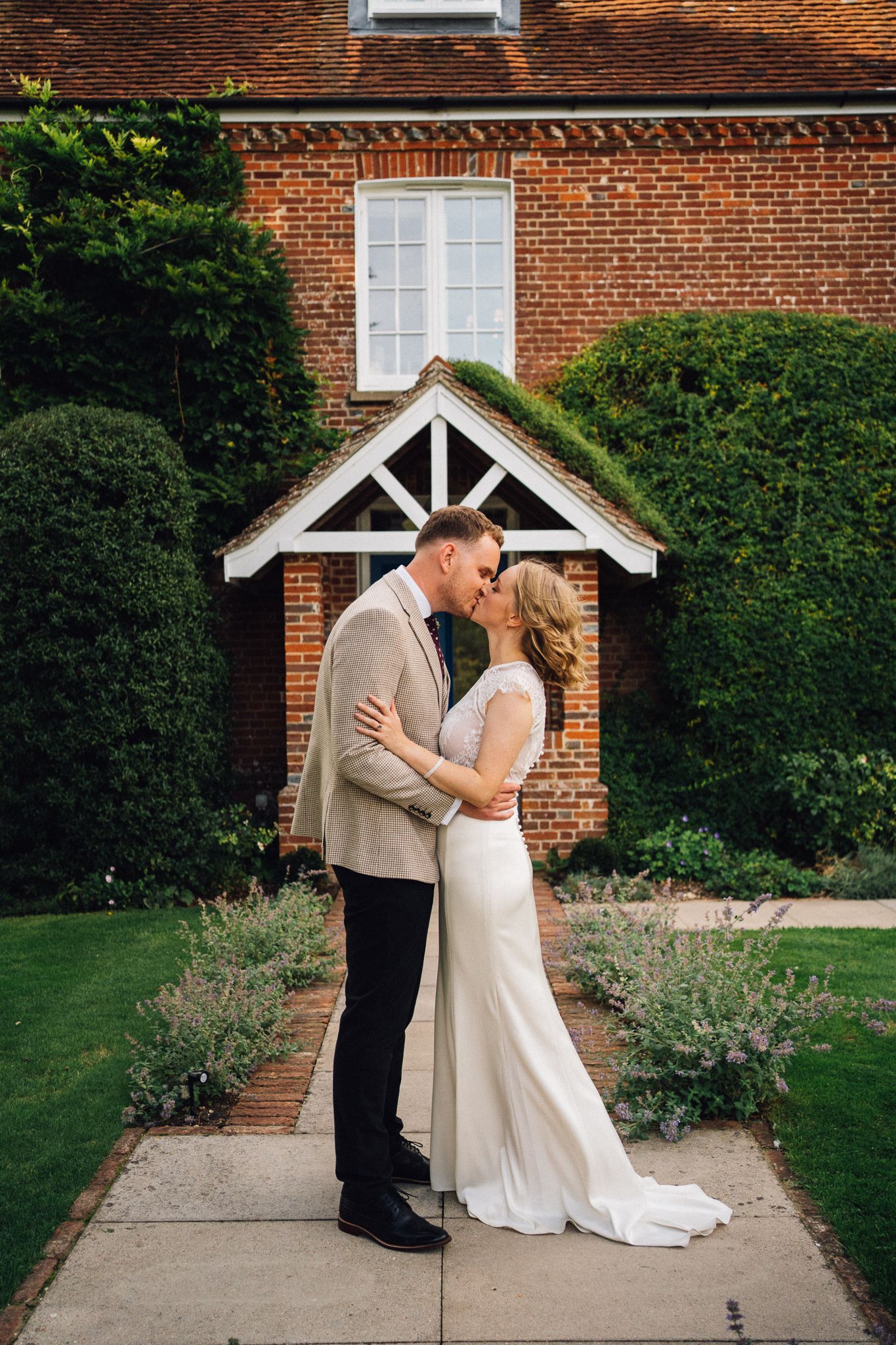 A couple shares a romantic moment under a brick entryway with white trim surrounded by manicured gardens.