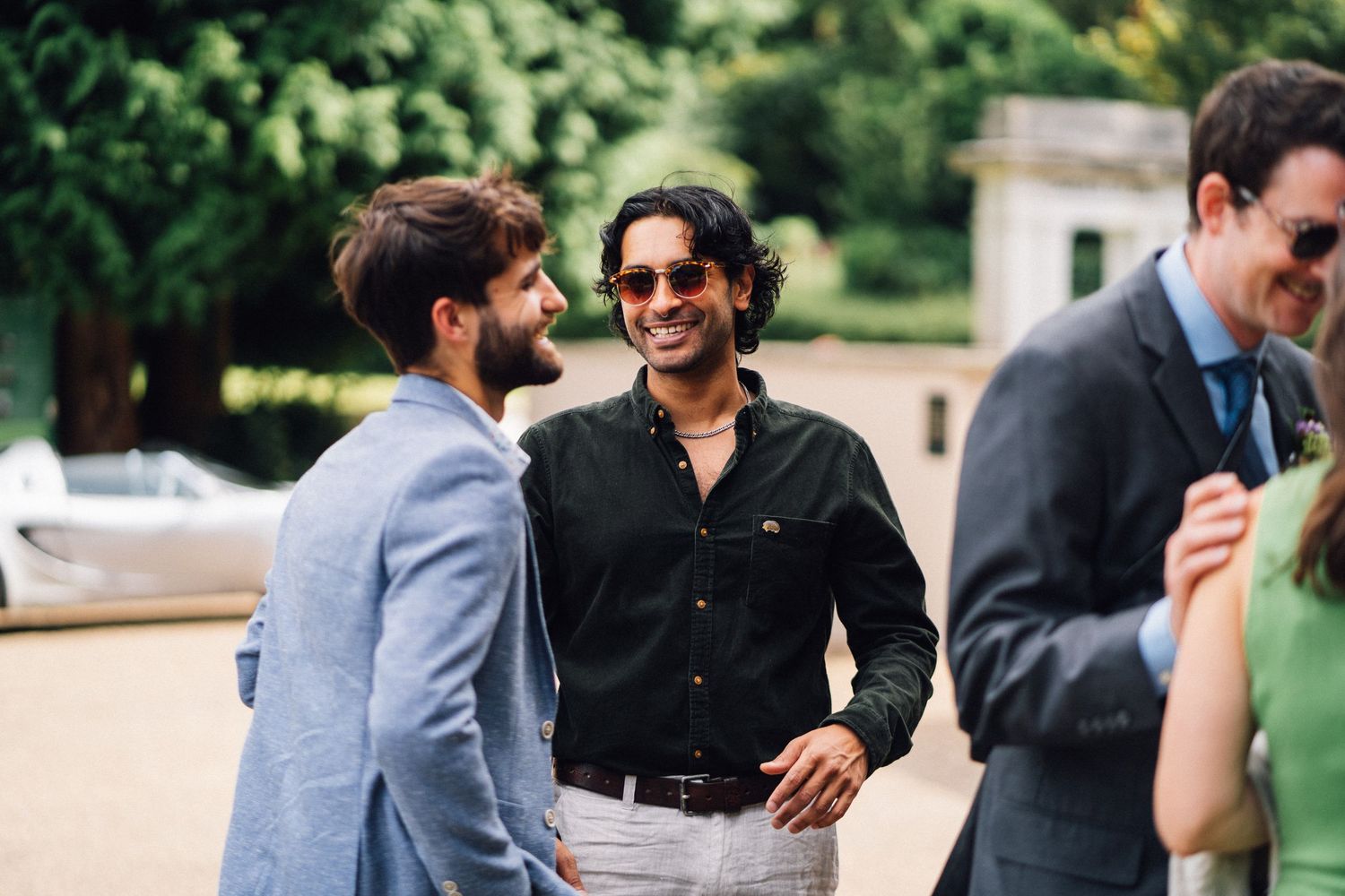 Wedding party members in suits and formal attire laugh and interact outdoors in natural lighting.