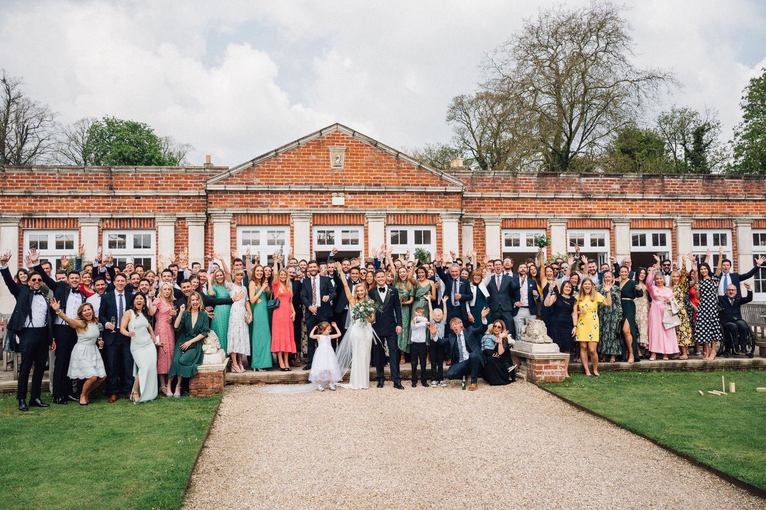 Wedding guests gather outdoors in formal attire for a panoramic group photo in front of a historic brick building.