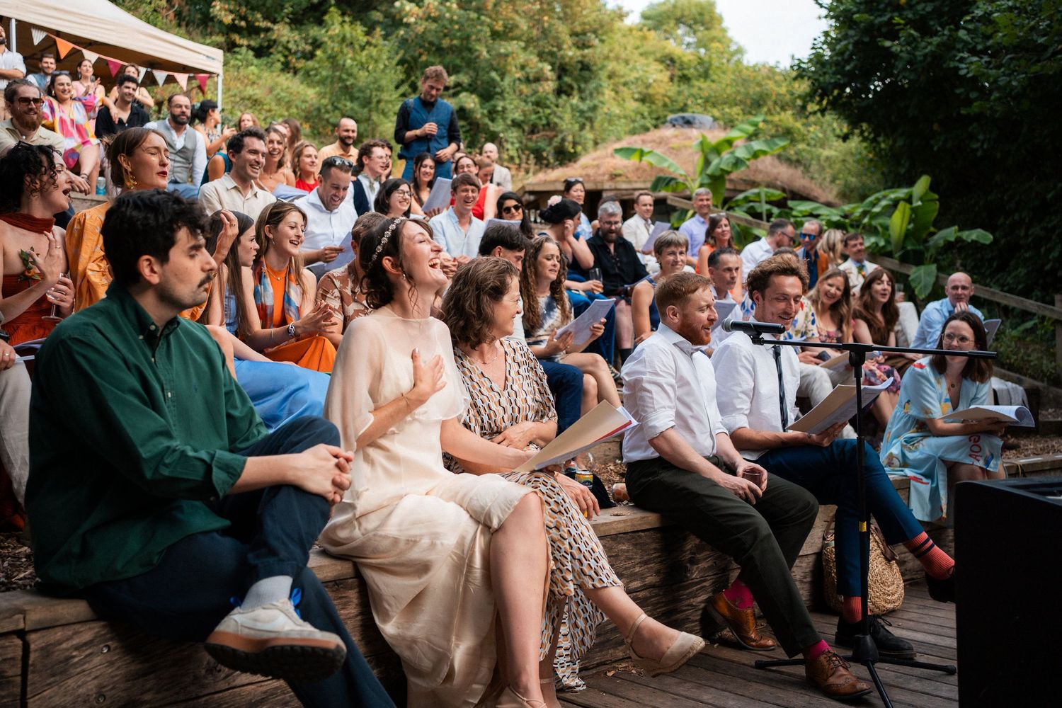 Crowd of guests laughing and enjoying an outdoor wedding ceremony seated on wooden steps surrounded by lush greenery.