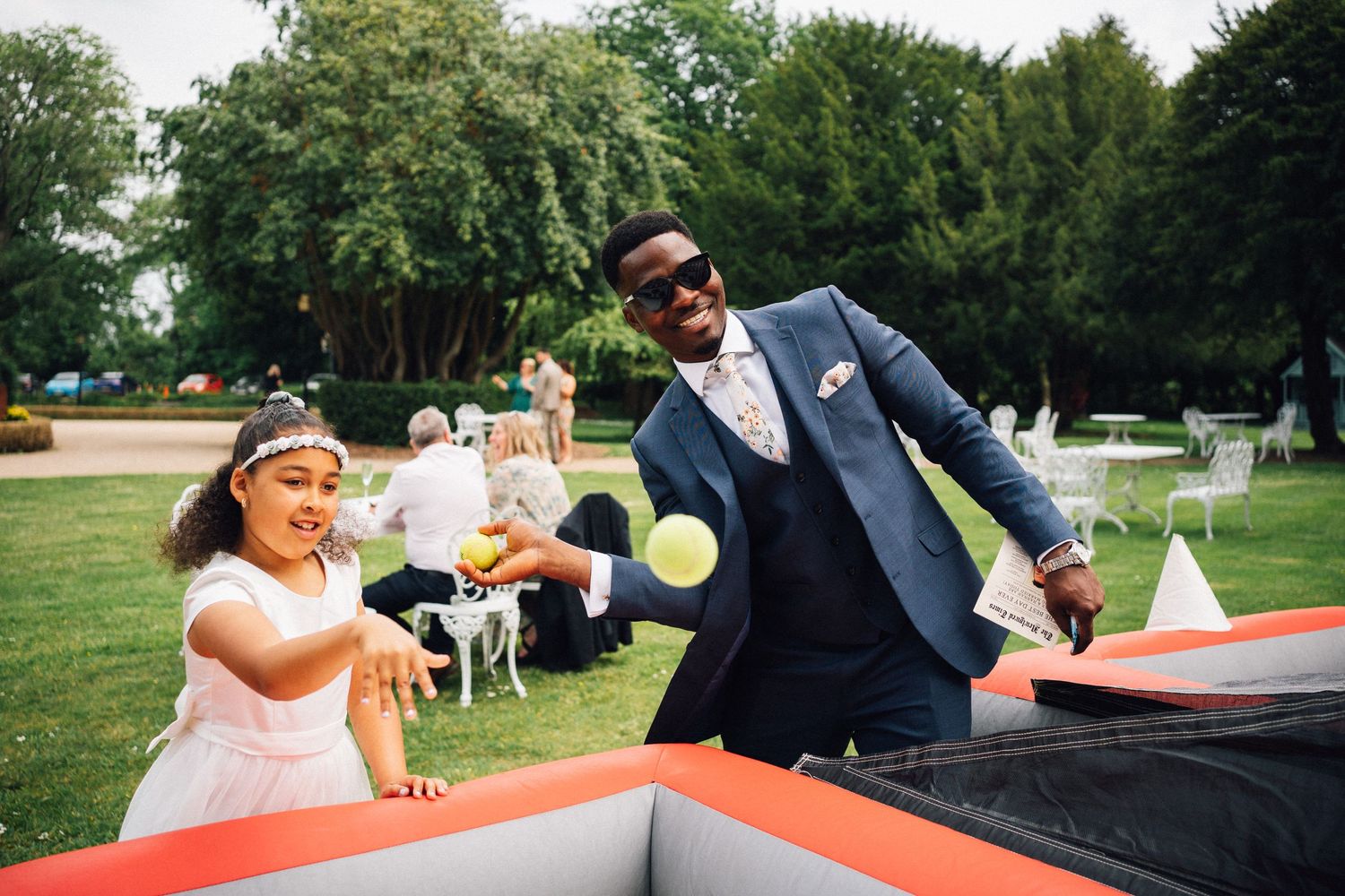 A man in a navy suit and a young girl in a white dress play beer pong together at an outdoor wedding reception.