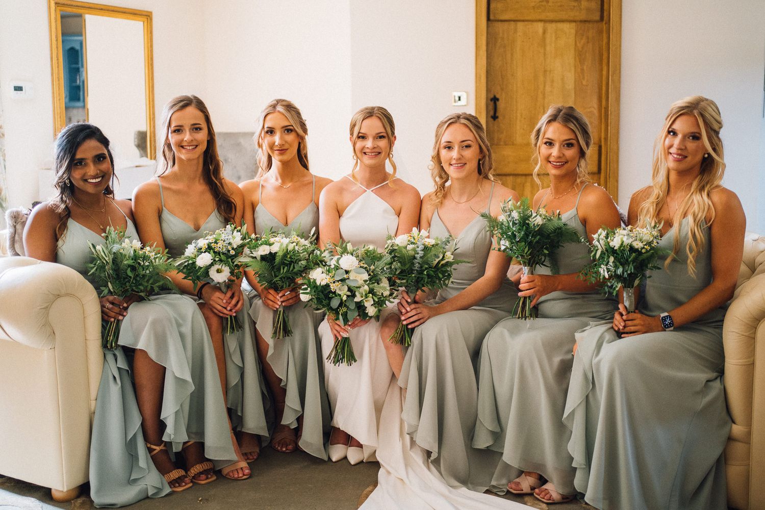 Bridesmaids in sage green dresses lined up indoors holding floral bouquets.
