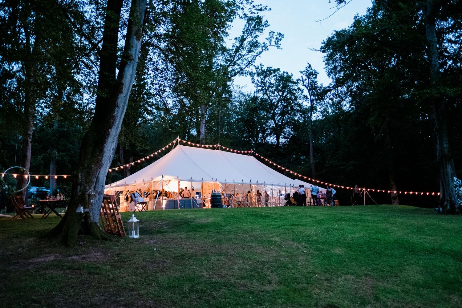 A glowing white wedding tent illuminates a grassy hillside at dusk with string lights.