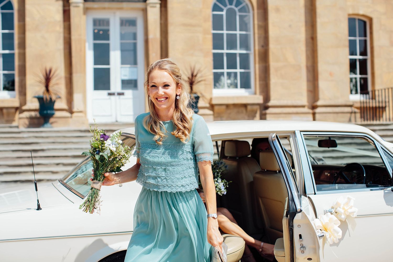 Wedding party members in light blue dresses gather outside a historic stone building.
