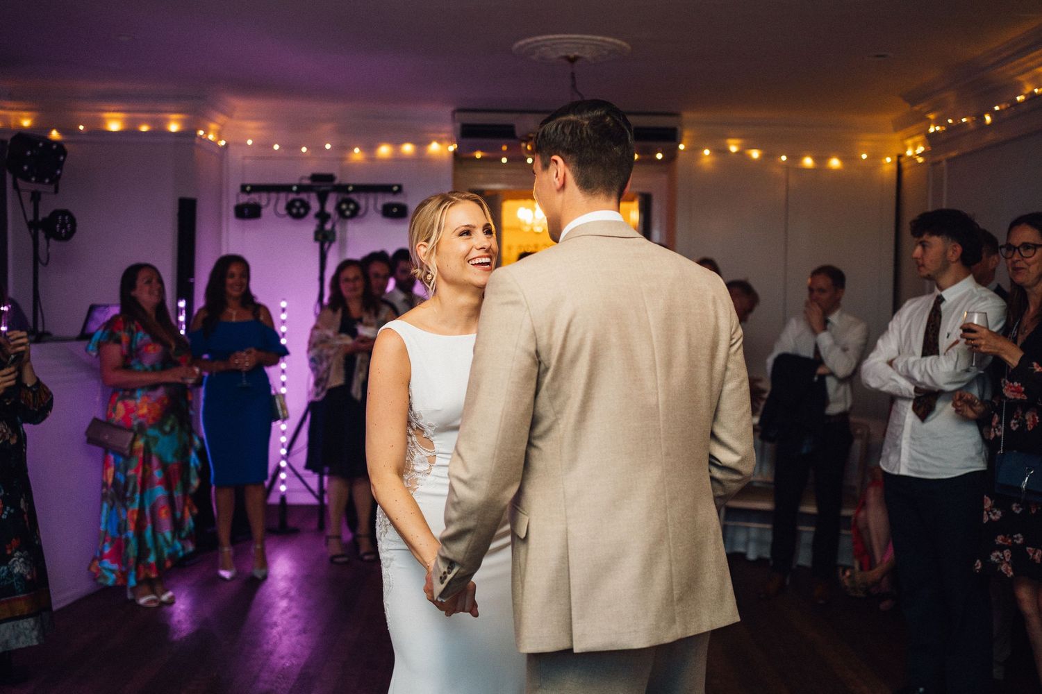Evening wedding reception with couples dancing under warm string lights and purple ambient lighting.