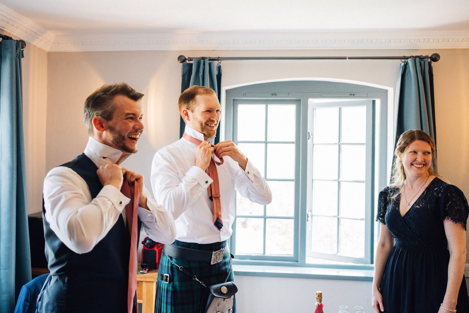 Groomsmen laughing and adjusting ties while preparing in a sunlit room with blue curtains.