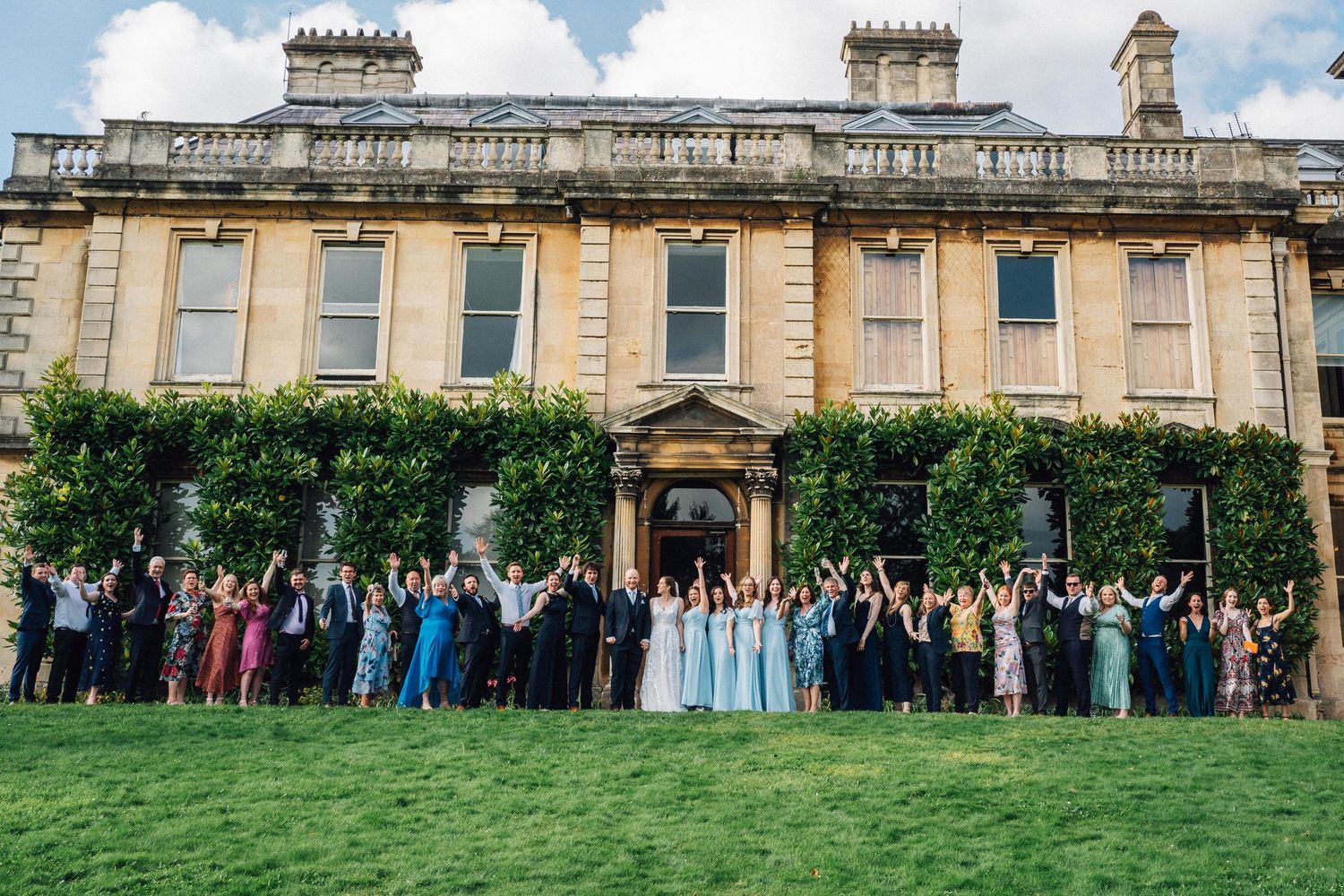 Large group of wedding guests gathered on a lawn in front of a grand historic building.