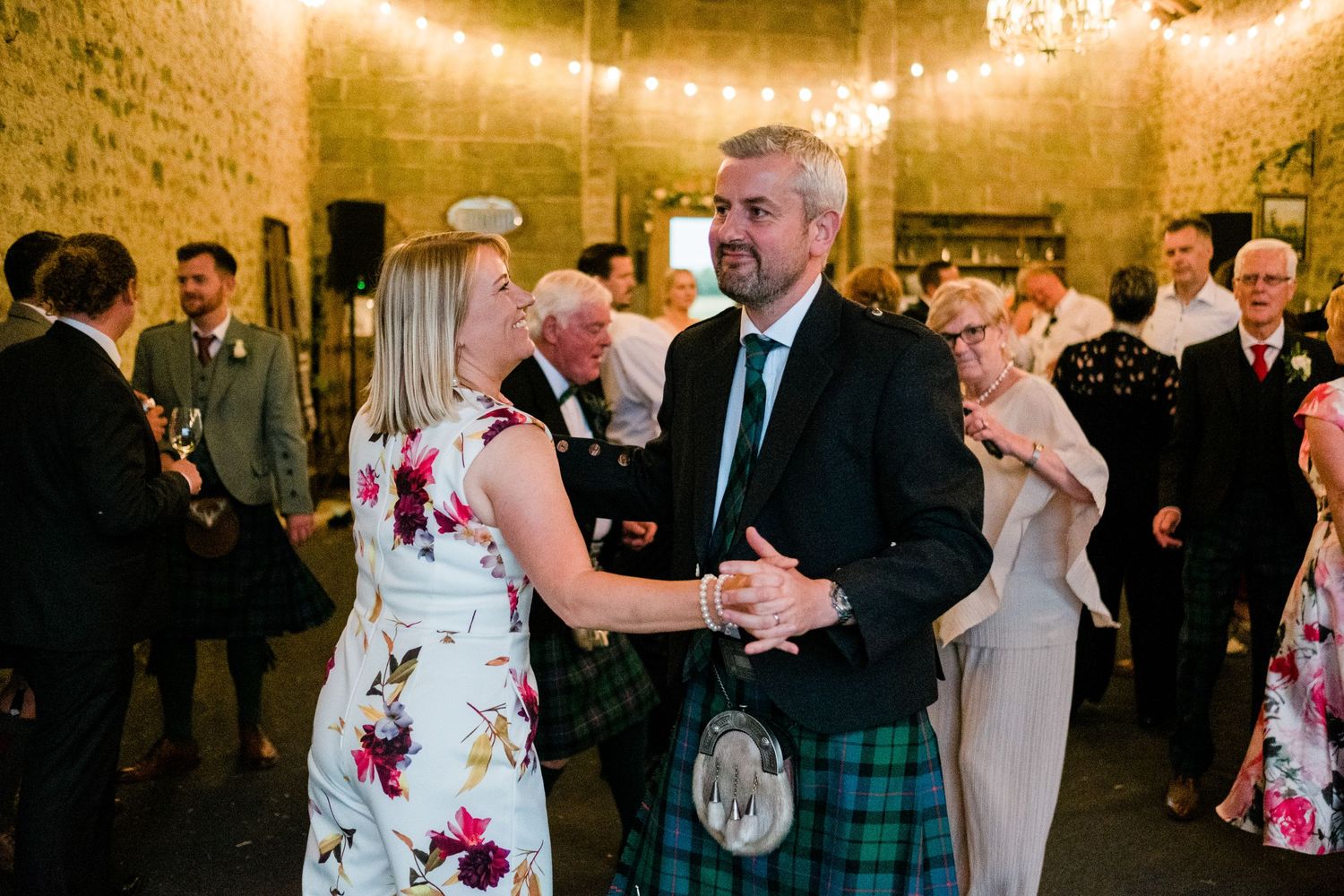Guests dancing at an elegant wedding reception in a rustic stone venue with string lights overhead.