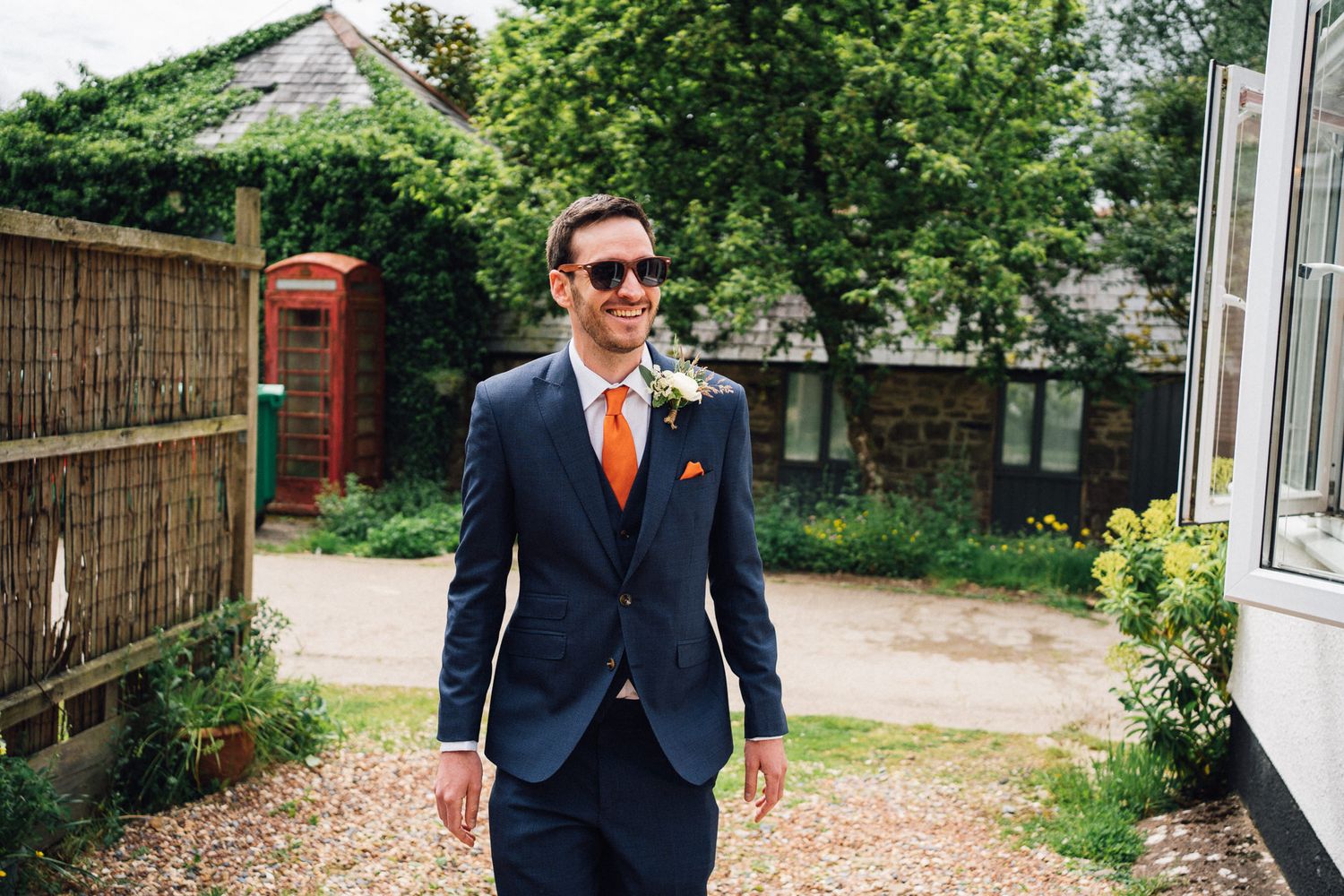 Wedding guests walk along a garden path between rustic buildings on a sunny day.