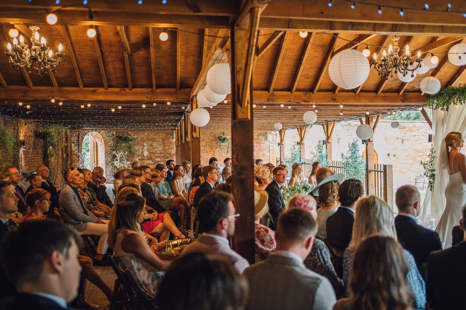 Wedding ceremony taking place in a rustic wooden barn venue decorated with chandeliers and white paper lanterns.