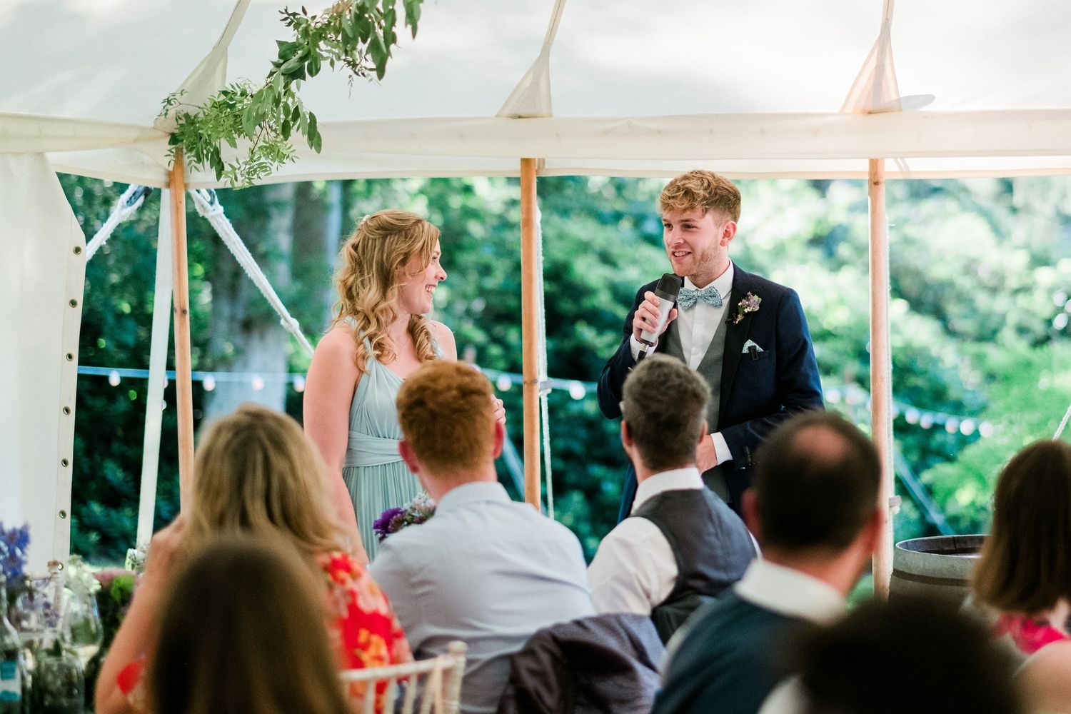 Wedding guests enjoy a lively reception under an elegant white tent decorated with greenery and floral arrangements.