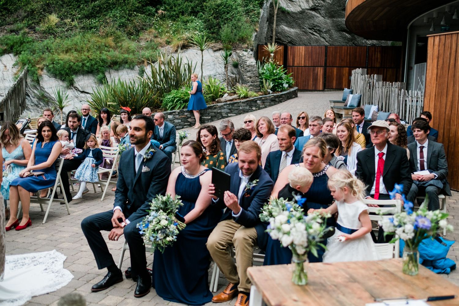 Outdoor wedding reception with guests seated on wooden benches surrounded by greenery.