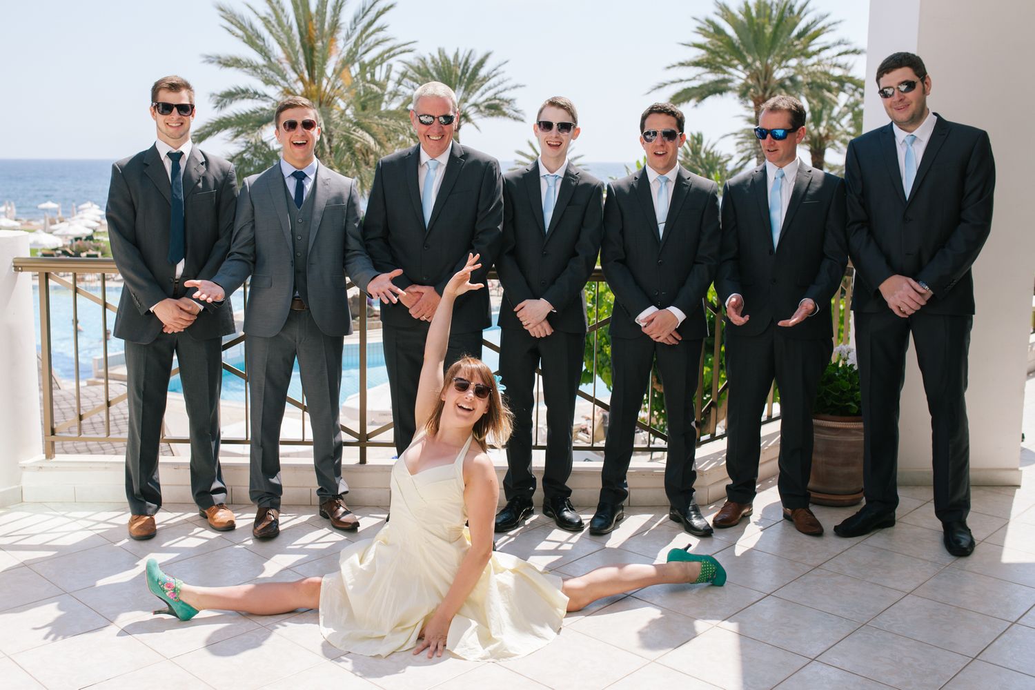 Wedding party in formal attire poses outdoors with palm trees and ocean views.