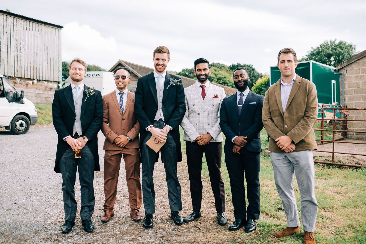 Wedding party groomsmen pose casually in front of rustic barn wearing mixed formal attire.