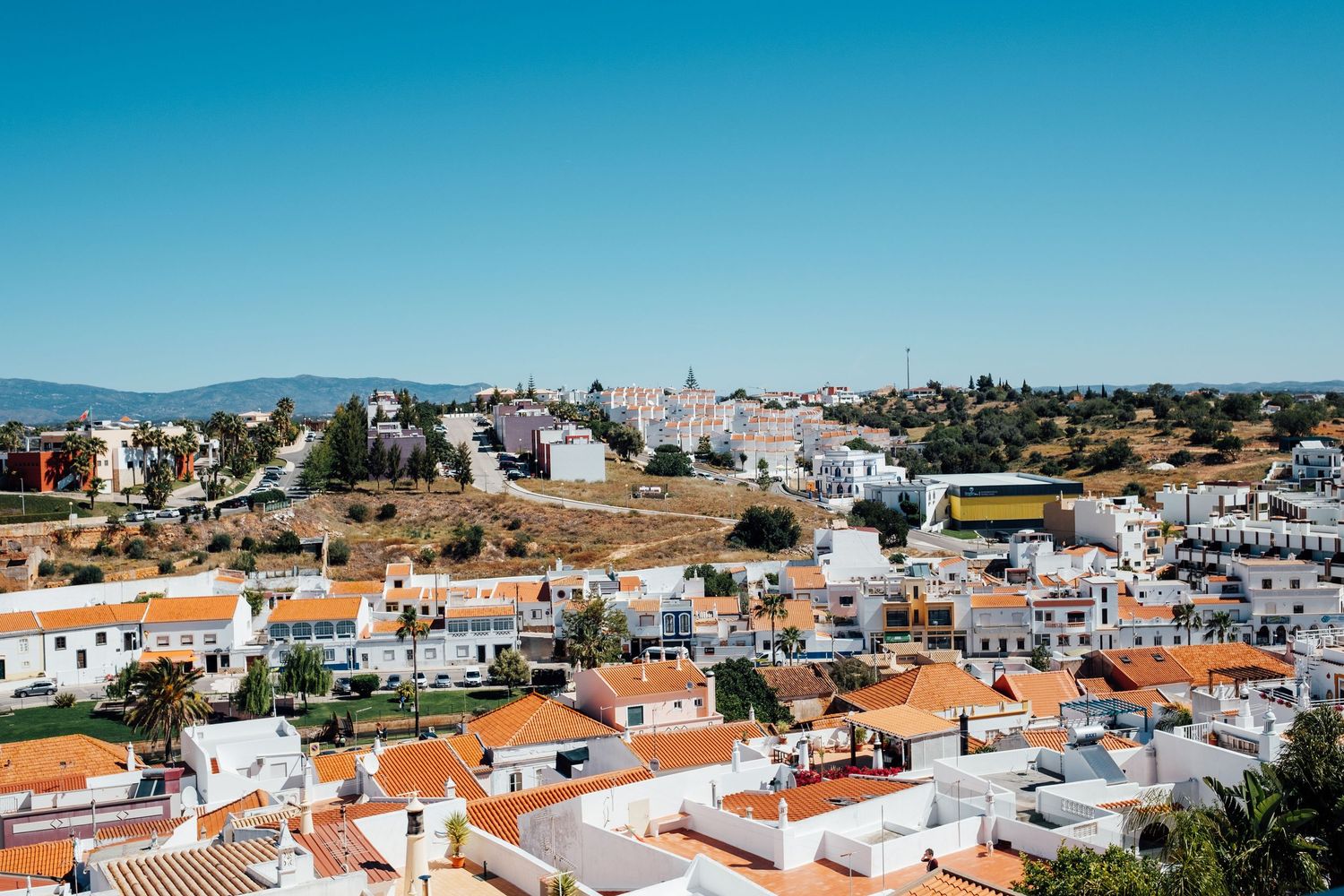 Panoramic view of white buildings with terracotta roofs in a coastal Mediterranean town against bright blue sky.