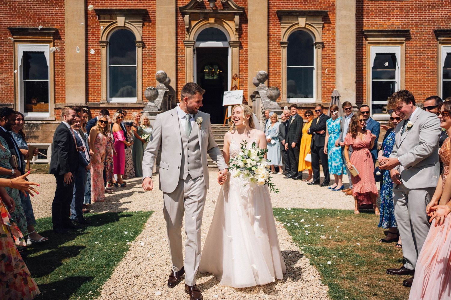 Wedding guests gather outside a brick building on a sunny day for an outdoor celebration.