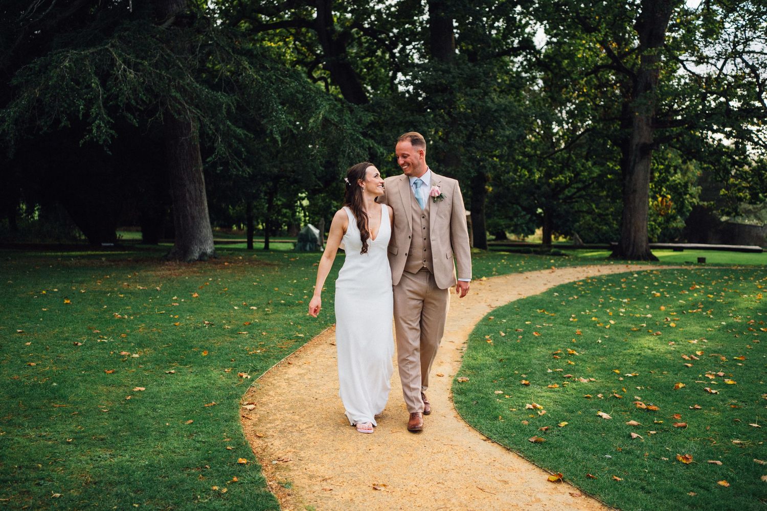 Newlyweds walk together along a winding garden path surrounded by green lawn and trees.