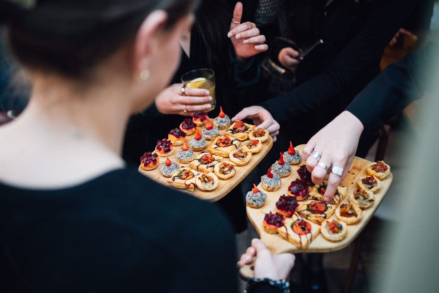 Hands holding wooden serving boards with assorted gourmet appetizers and hors d'oeuvres at an elegant event.