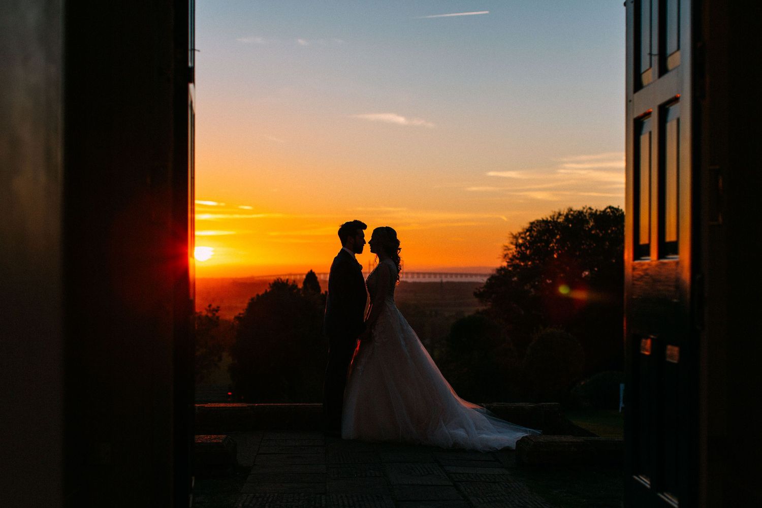 Silhouettes of a couple stand against a vibrant orange sunset sky framed by architectural elements.