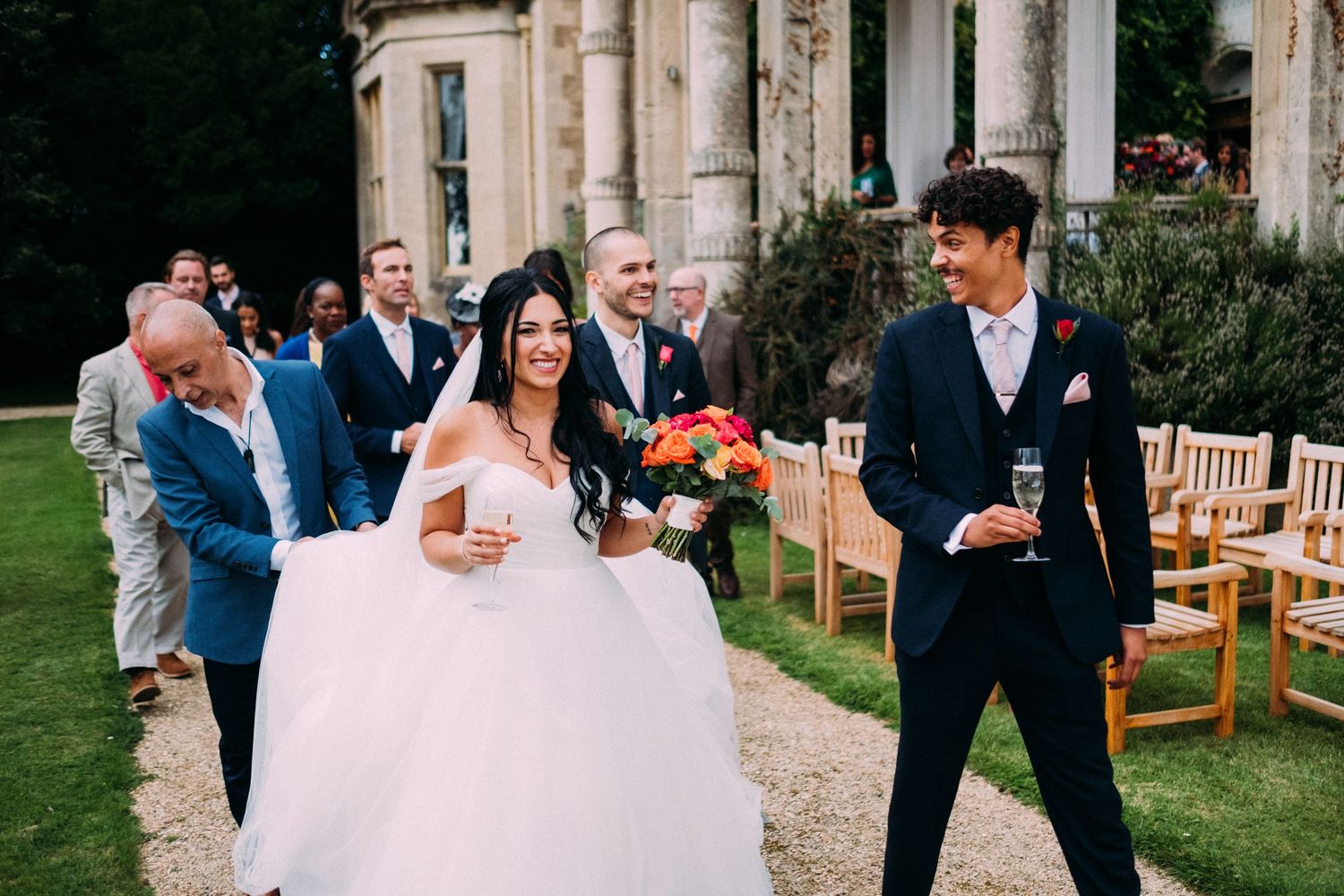 Wedding guests mingle and celebrate during an outdoor garden reception with wooden furniture and floral arrangements.