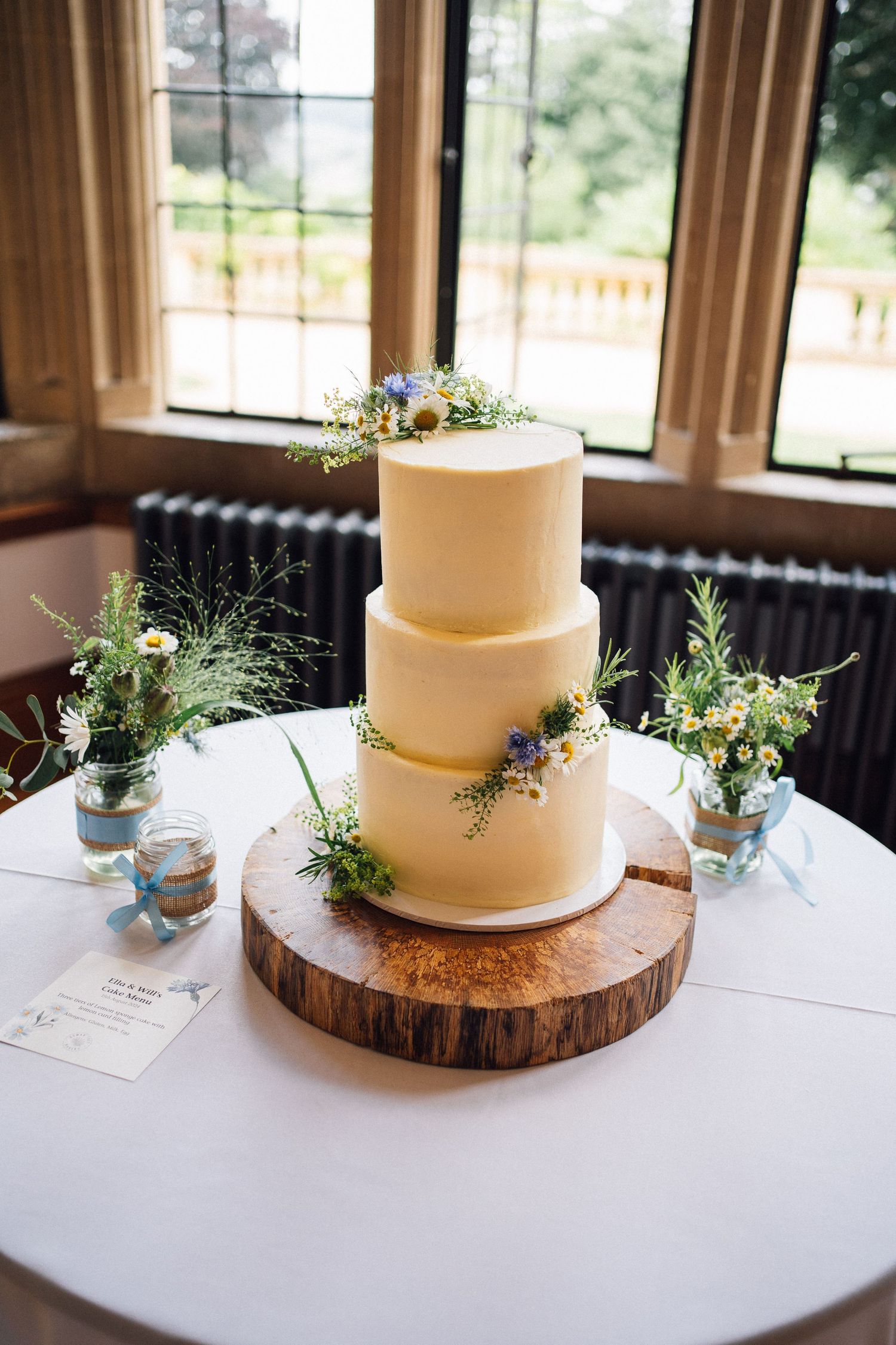 Simple white wedding cake with fresh greenery displayed on rustic wood slice at elegant indoor reception venue.