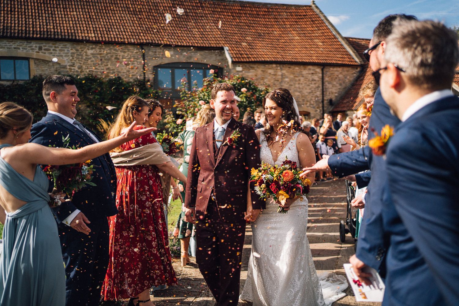 Wedding guests throw confetti over a couple outside a rustic brick building during a joyful celebration.