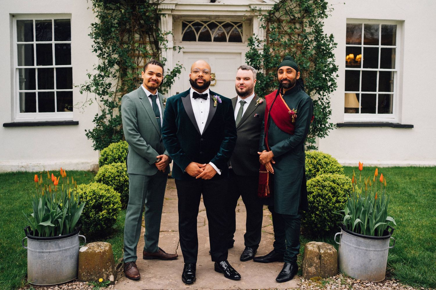 Groomsmen in black tuxedos and bowties gather outside a white building with elegant windows and potted plants.