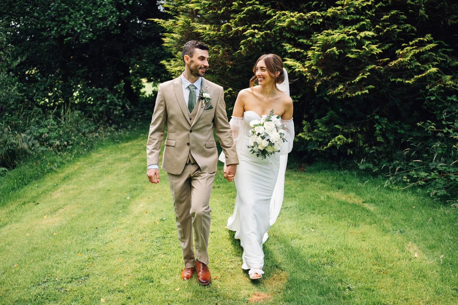 Wedding couple and guests walking through lush garden path during outdoor summer celebration.