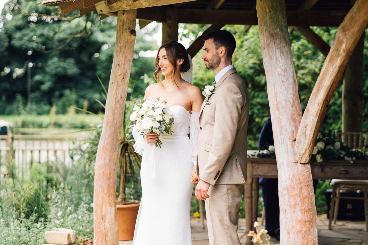 A couple shares an intimate moment under a rustic wooden pergola surrounded by lush garden scenery.