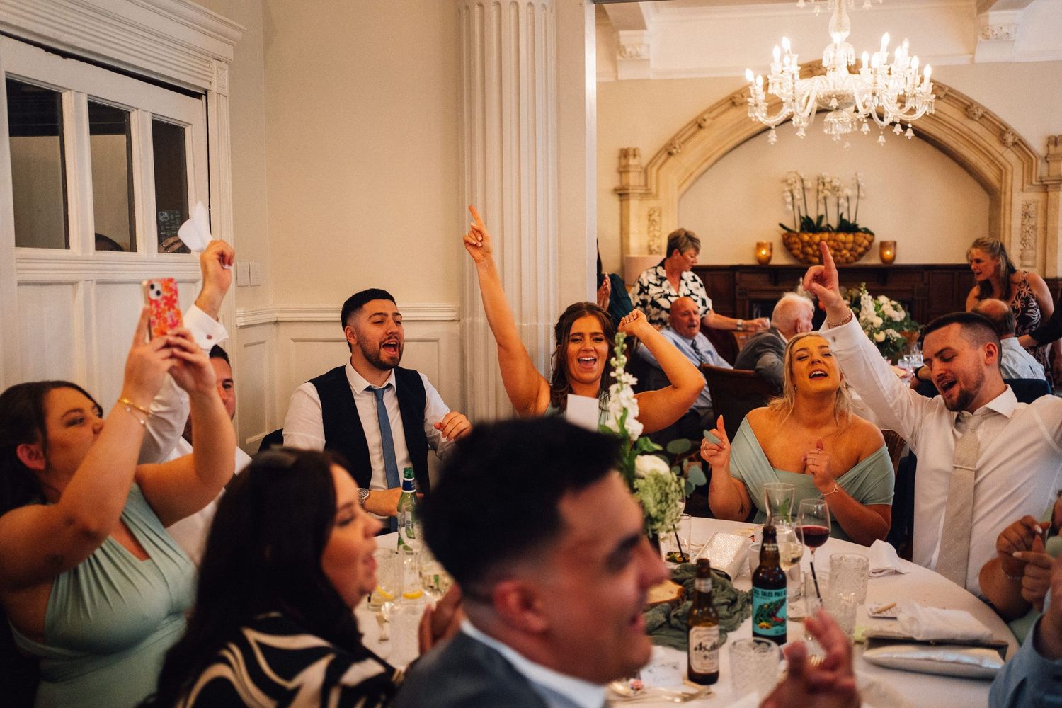 Wedding reception guests celebrate and dance together in an elegant white-walled venue with chandeliers.