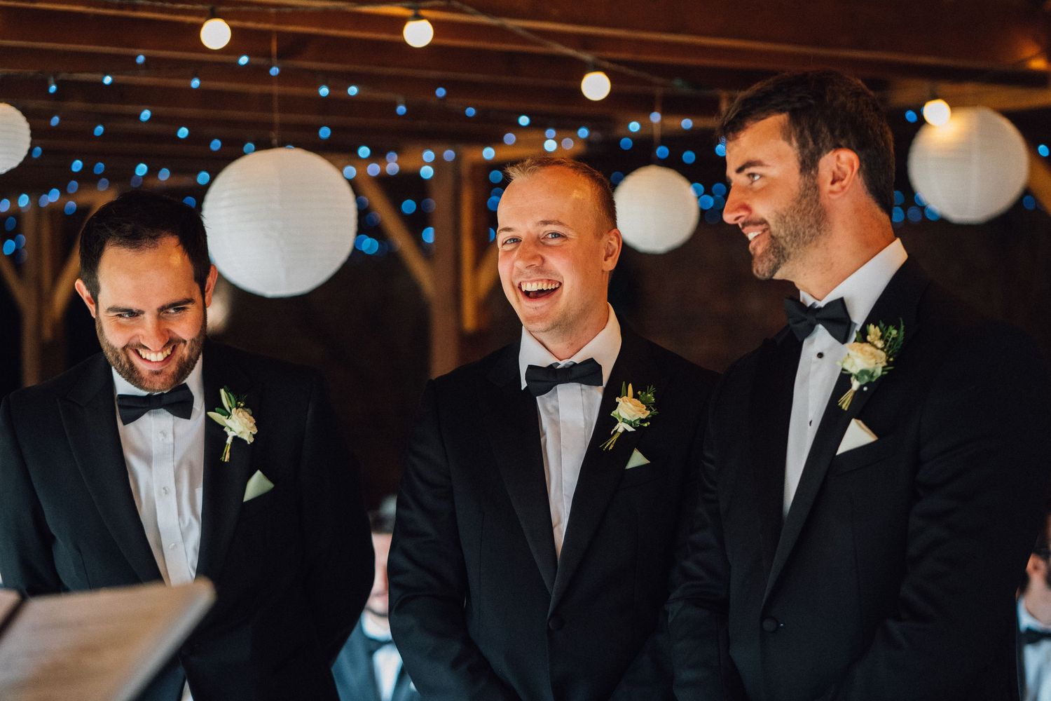 Groomsmen share laughter and smiles during wedding reception under white paper lantern decorations.