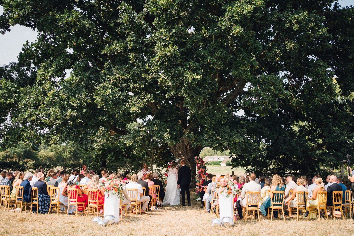A panoramic view of people seated in wooden chairs outdoors under a canopy of trees during a gathering.