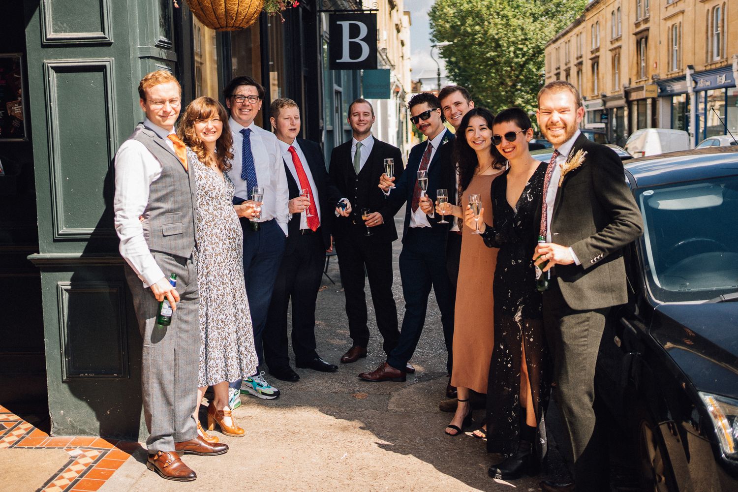 A group of well-dressed guests gather outside on a sunny day in front of a brick building.