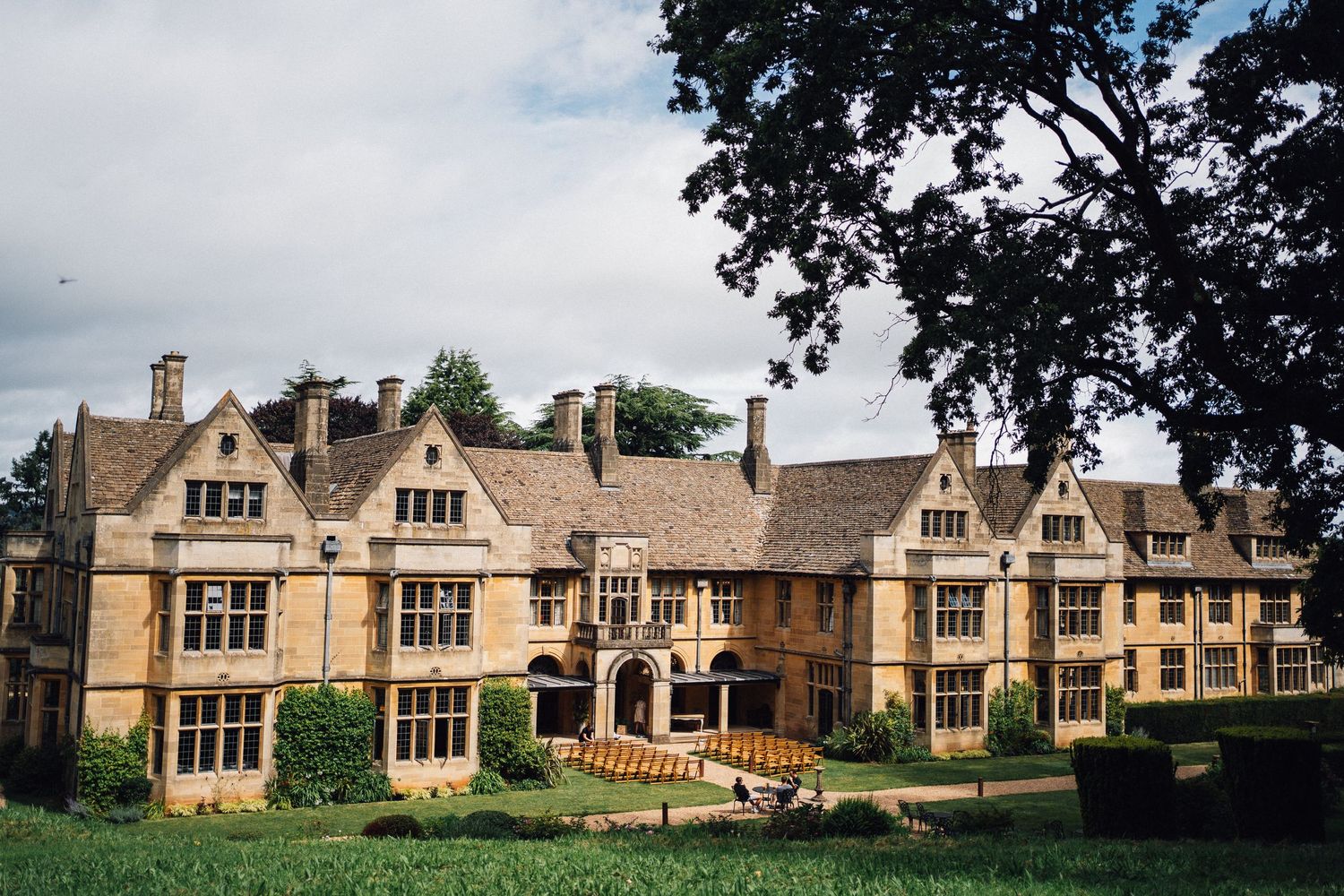 Historic English manor house with honey-colored stone facade and ornate Tudor-style architecture set on manicured grounds.