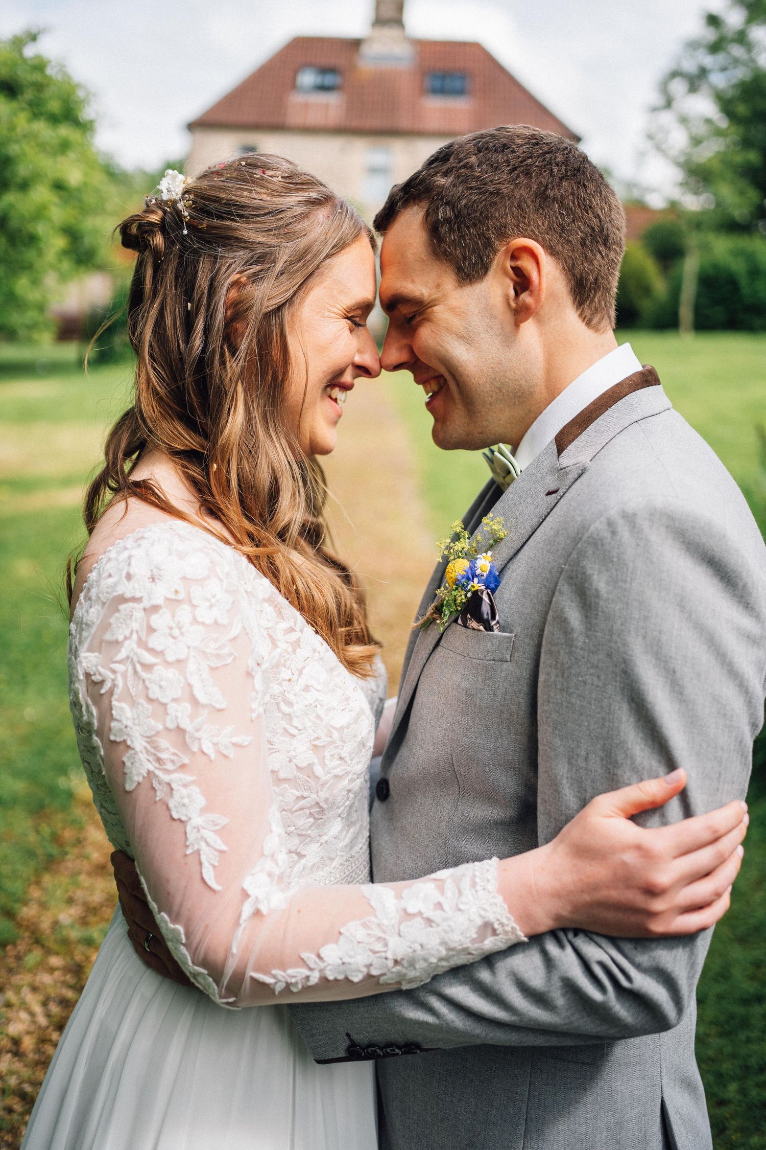 Romantic wedding couple embraces in a garden setting wearing a lace sleeve dress and grey suit.