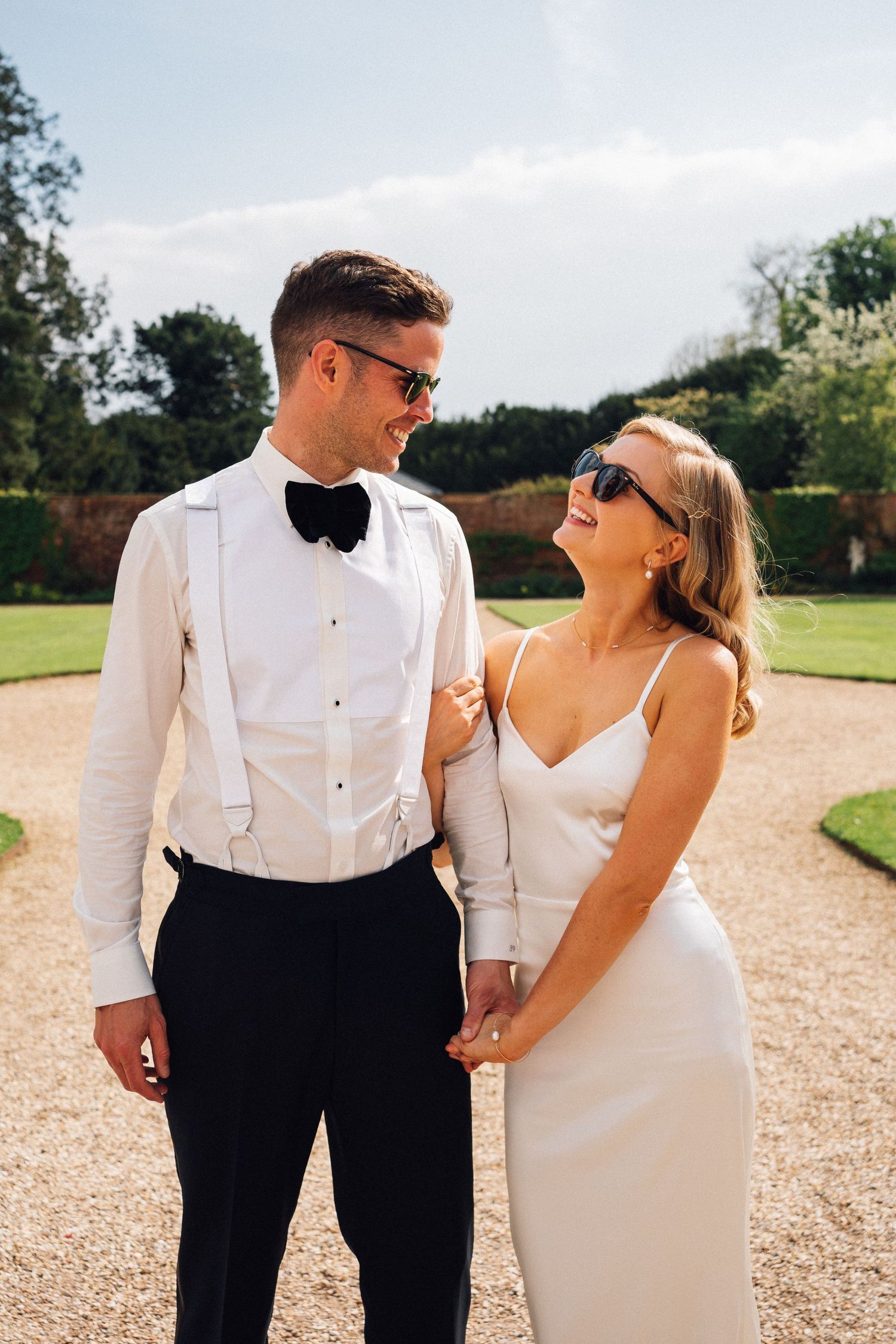 Couple in formal wedding attire walking together and laughing on a sunny outdoor path.