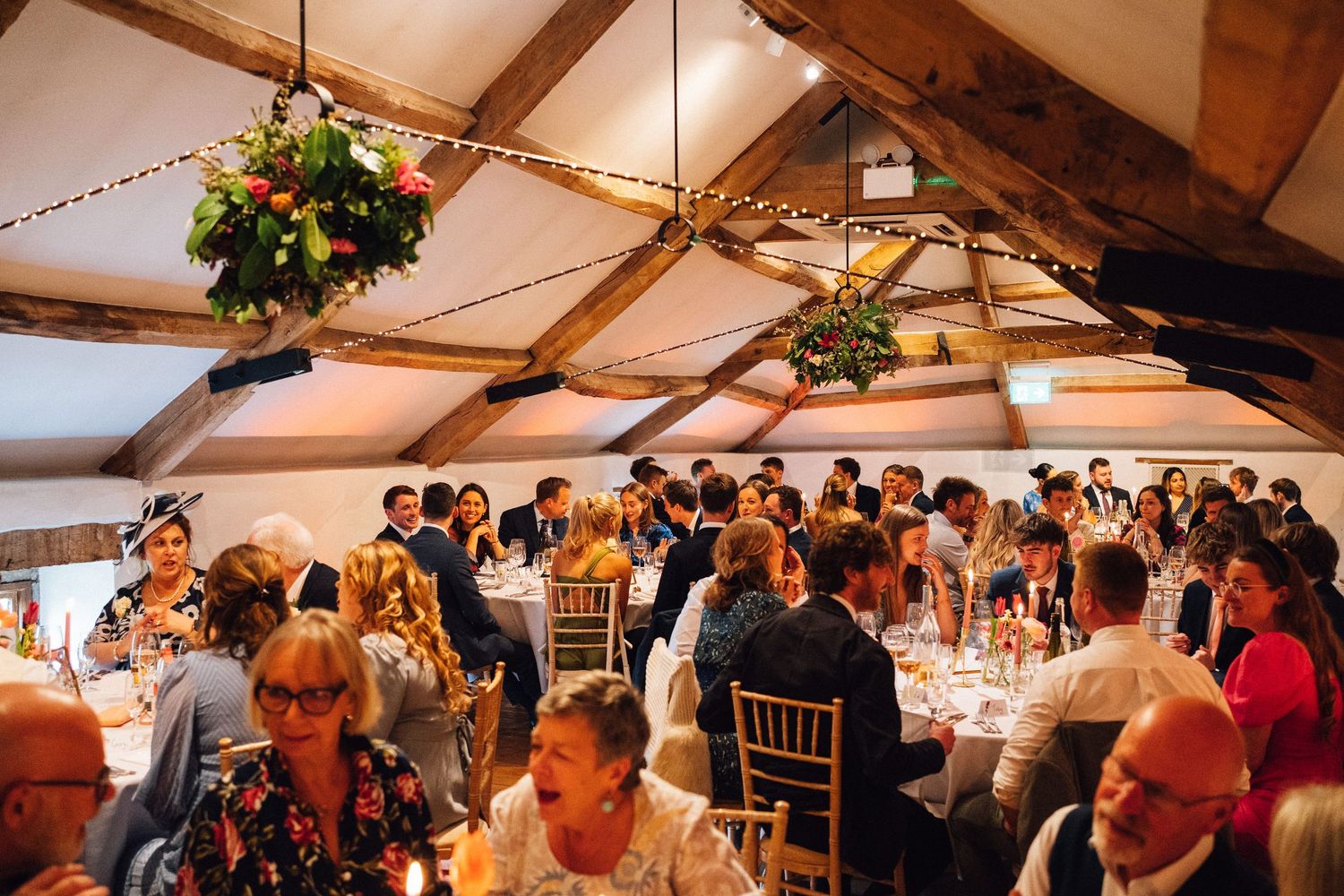 Wedding reception venue with exposed wooden beams and hanging floral decorations during dinner service.