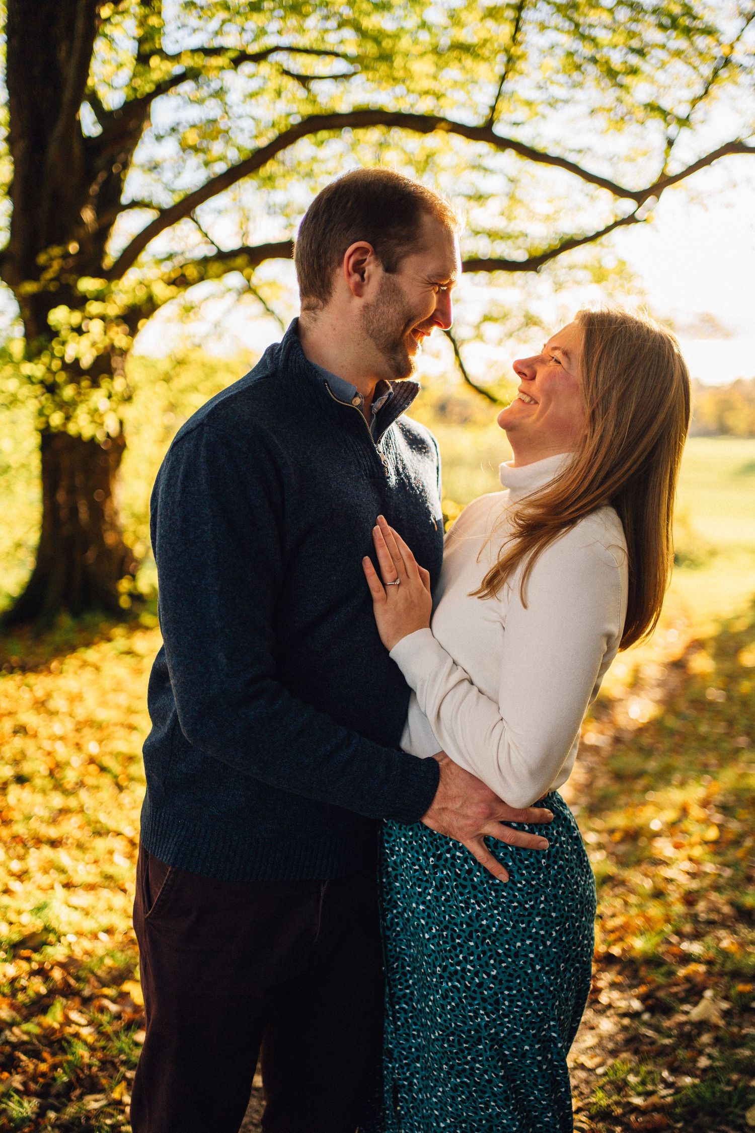 A couple embraces in golden autumn sunlight beneath trees with fallen leaves.