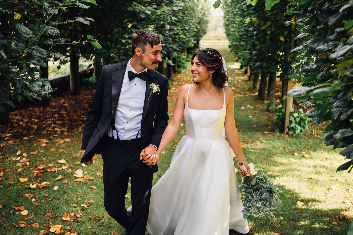 Couple in wedding attire walking together through a sunlit garden path.