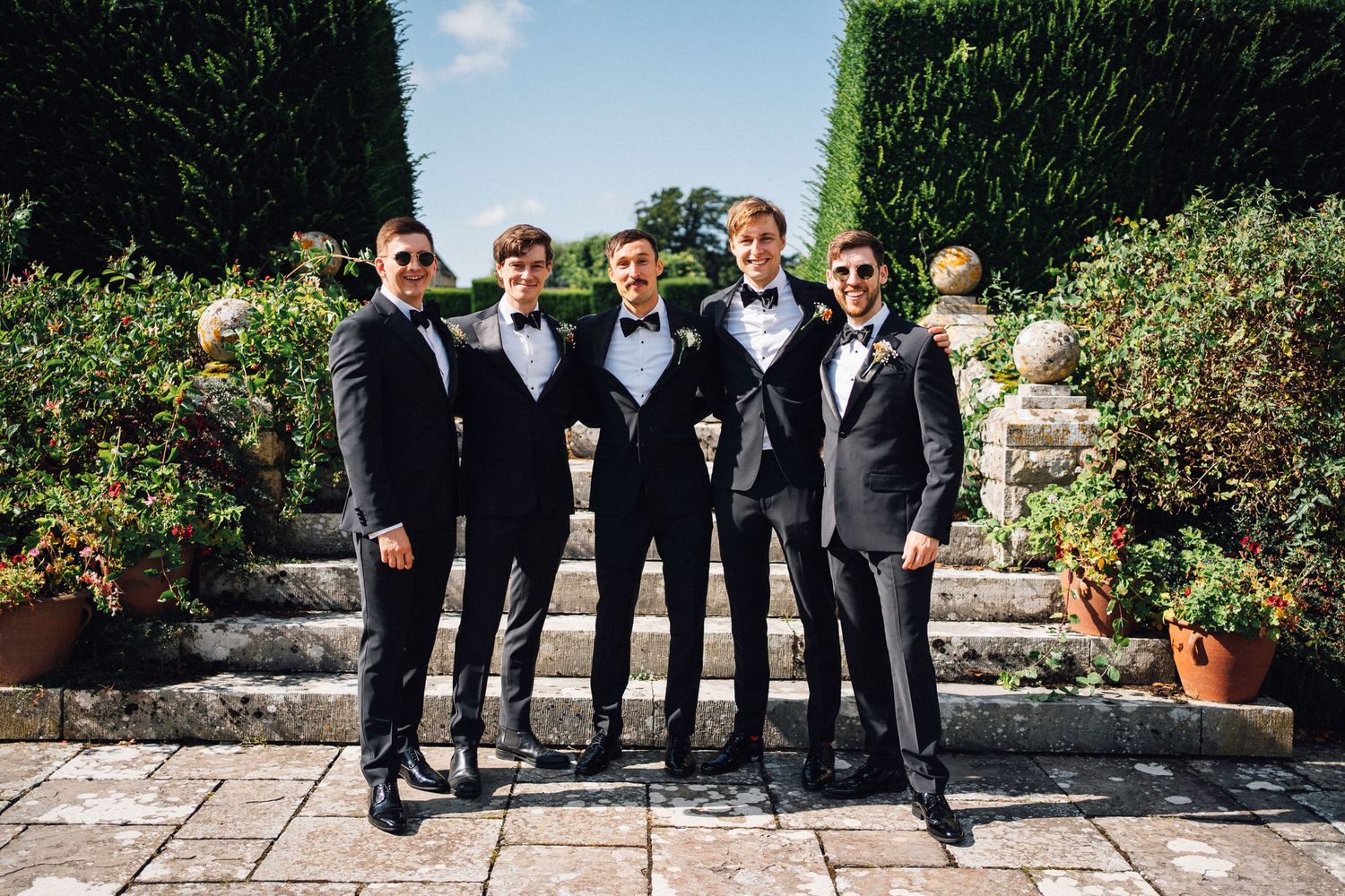 Groomsmen in black tuxedos pose together in a manicured garden setting.