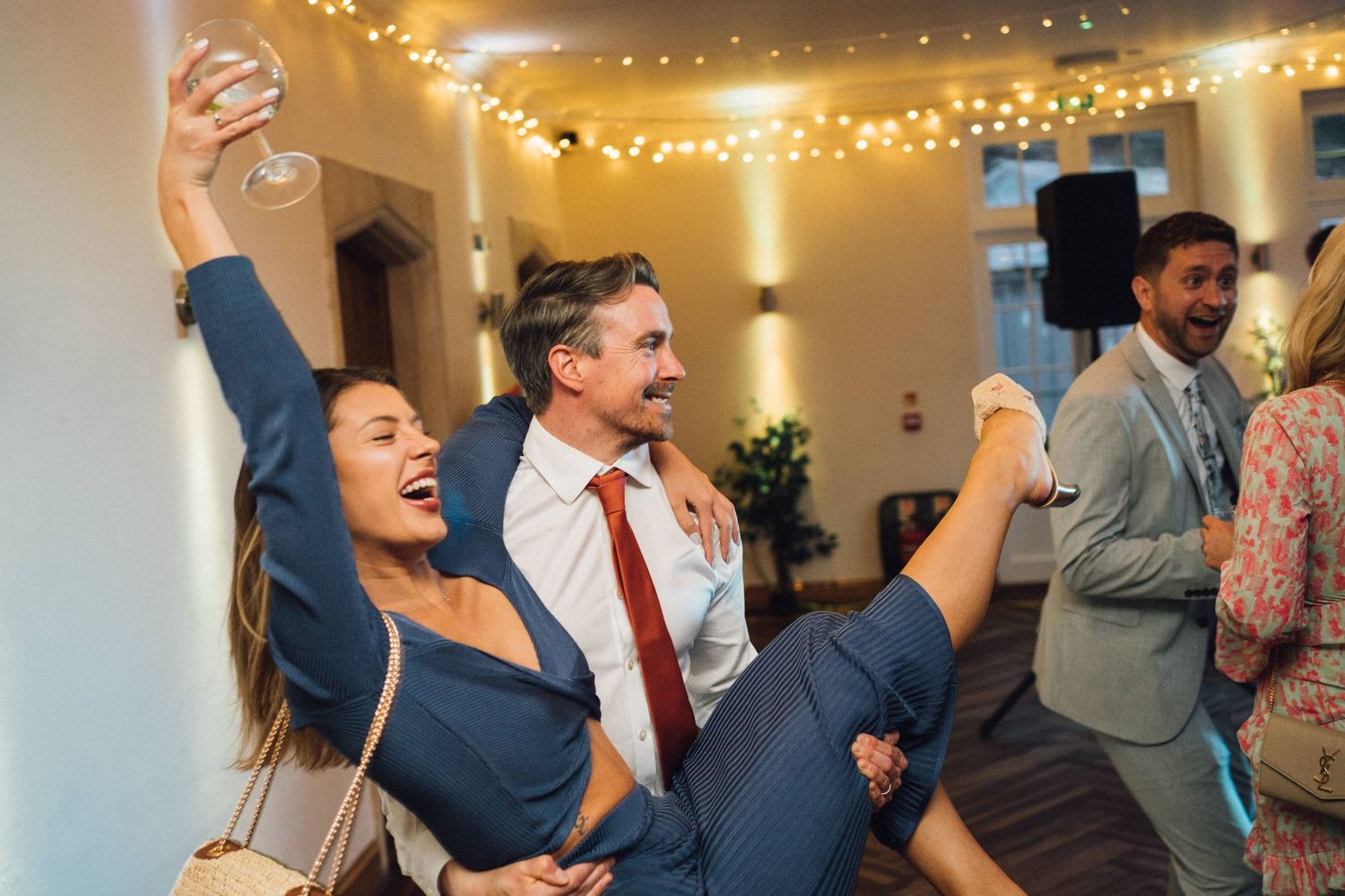 A joyful woman in a blue suit is lifted by a man at a lively wedding reception with fairy lights.
