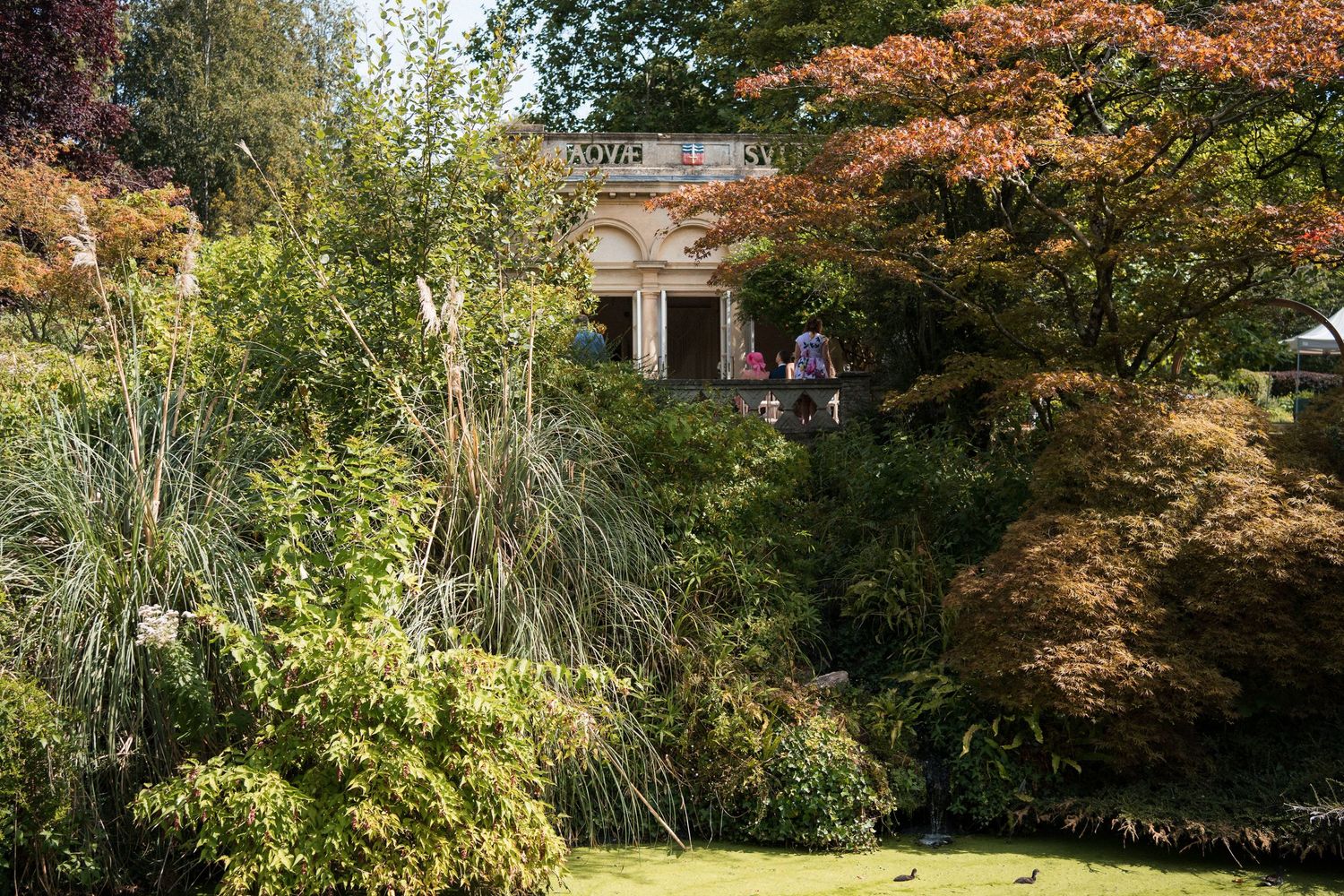 Lush garden with colorful autumn trees surrounding a classical stone building with visitors exploring the grounds.
