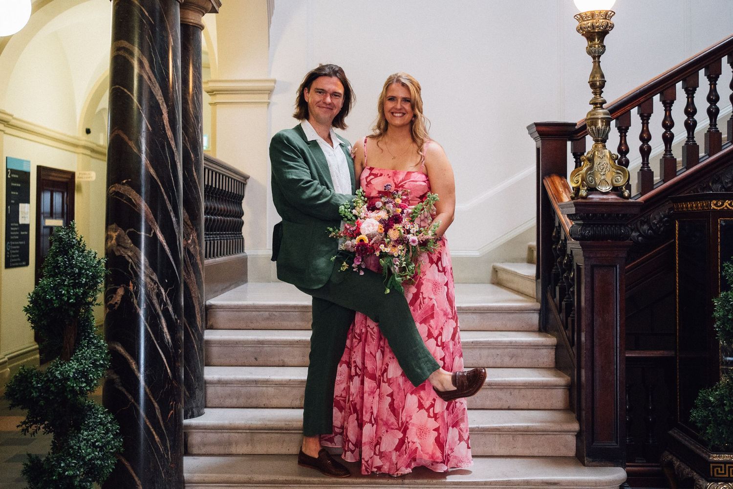 Couple smiling on marble staircase, man in green suit, woman in pink floral dress holding bouquet, elegant venue.