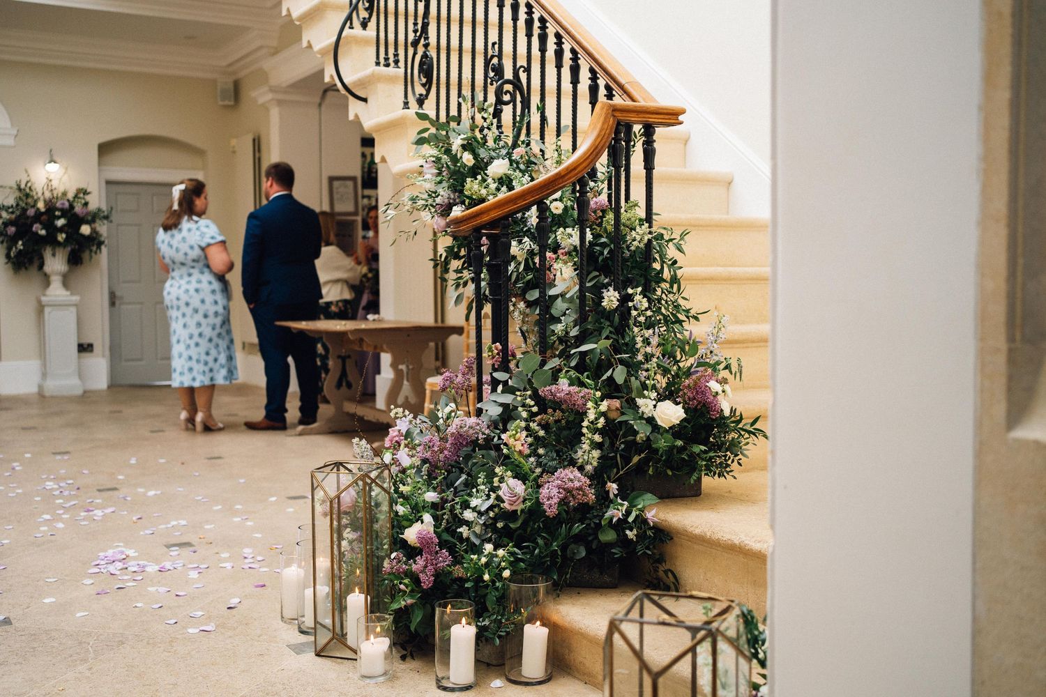 Grand staircase decorated with floral arrangements and white rose petals scattered on carpeted steps.