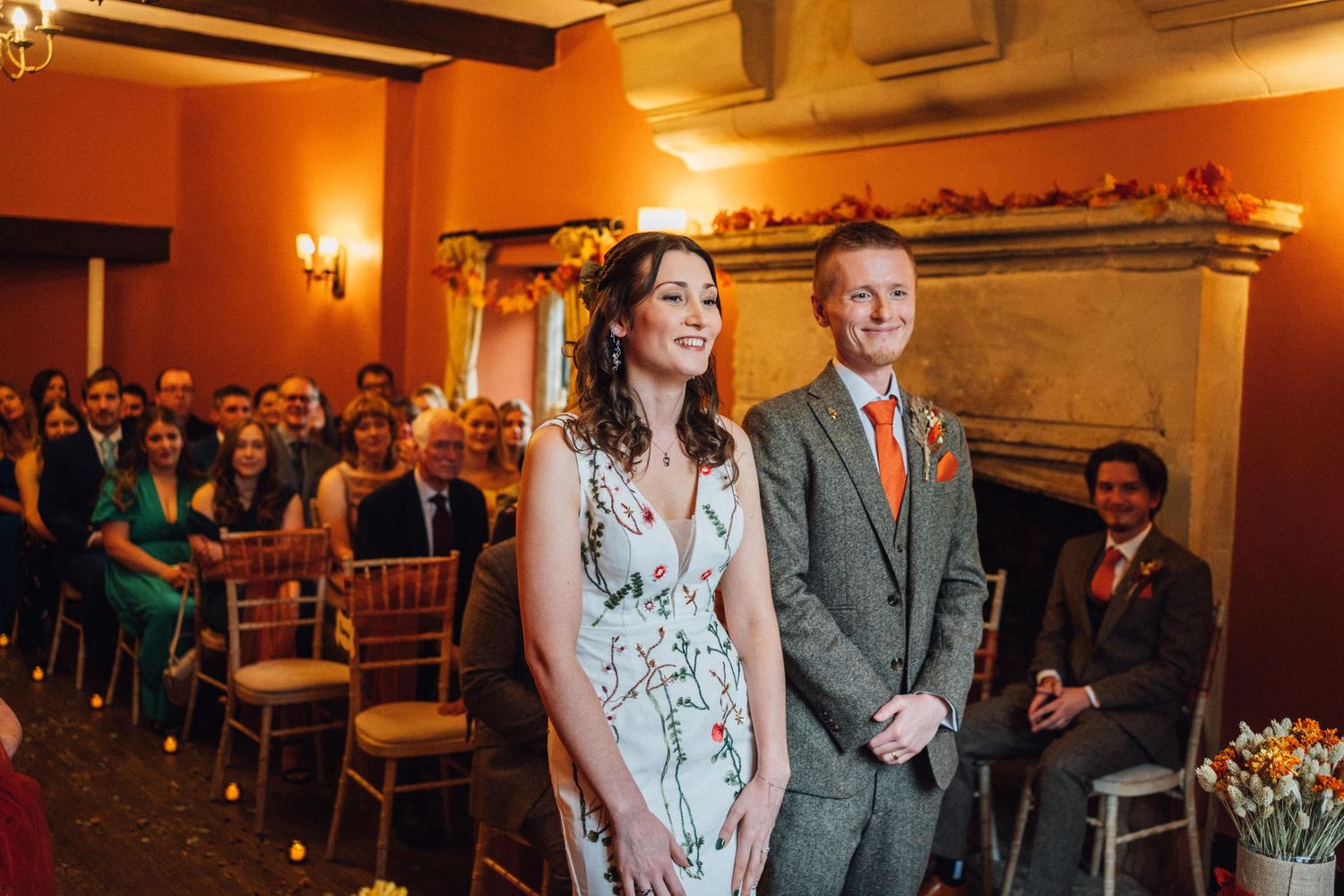 Wedding ceremony in a warm-lit room with orange walls and wooden beams, with attendees seated in rows of chairs.